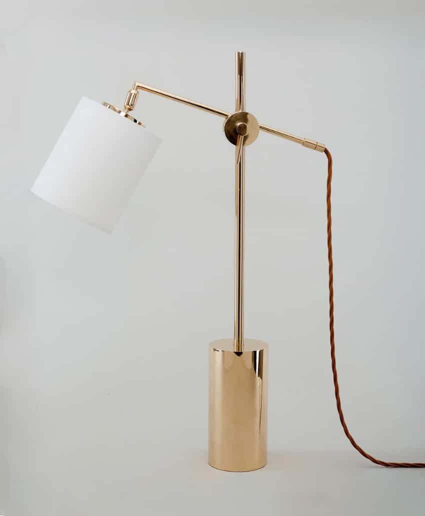 A modern table lamp with a gold metal base and stand, a cylindrical white lampshade angled to the side, and a brown twisted cord against a plain light background.