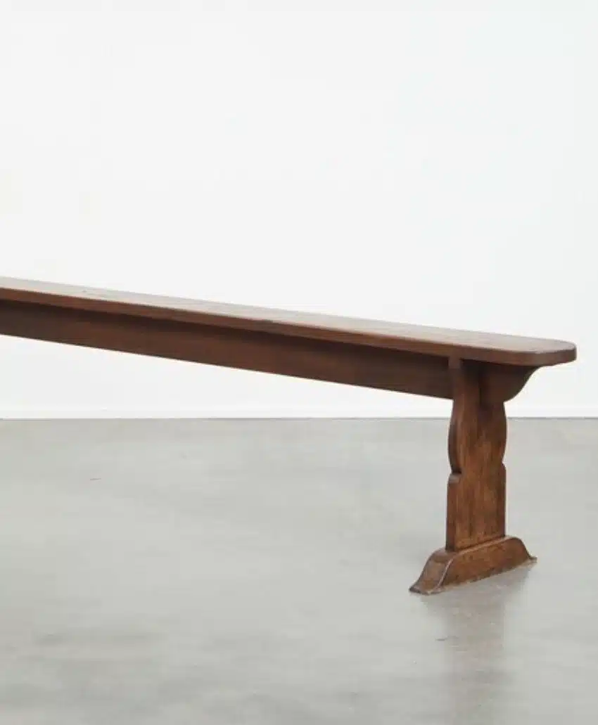 A long wooden bench with a simple design and sturdy legs sits on a smooth, gray concrete floor against a plain white wall.