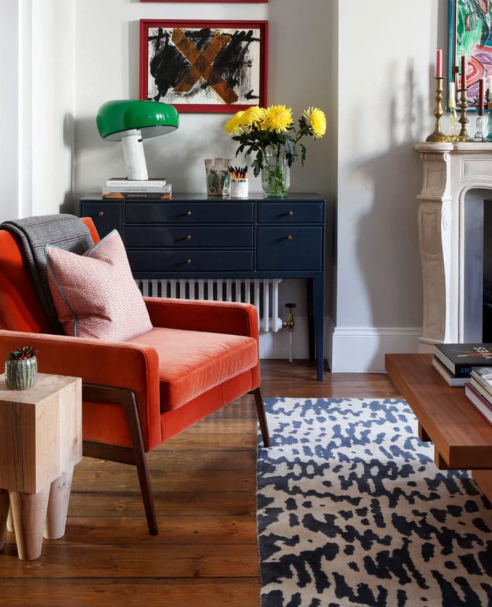 A cozy living room with an orange armchair, patterned rug, wooden coffee table, and side table. A dark blue console holds a green lamp, yellow flowers, and artwork. A white fireplace and candles are visible on the right.
