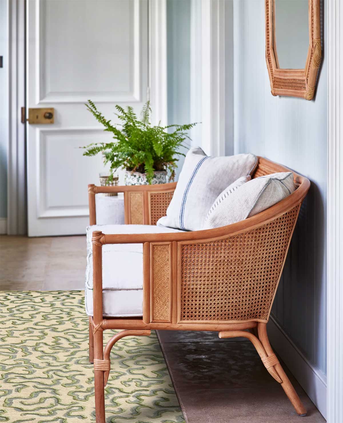 A rattan armchair with white cushions and striped pillows sits by a pale blue wall, with a potted fern on an end table and a patterned green rug on the floor. A mirror hangs above the chair near a white door.