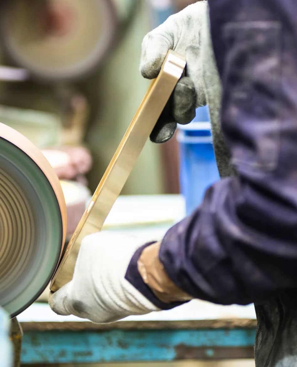 A person wearing gloves is polishing a metal object using a grinding or buffing wheel in a workshop setting.
