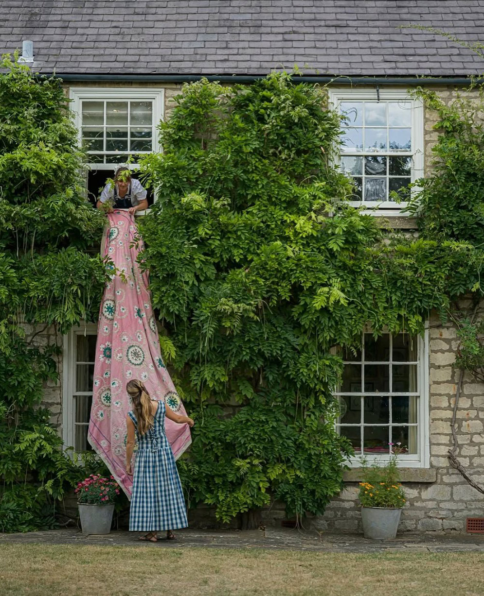 A woman in a checkered dress stands outside a window of a vine-covered house while another woman inside lowers a pink floral blanket for her to climb.