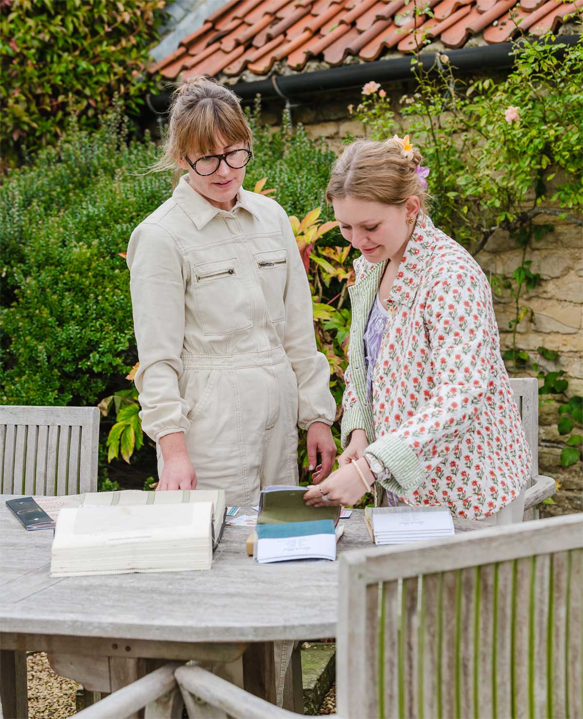 Two women stand at a wooden outdoor table with open books, engaged in discussion. One woman is wearing glasses and a light jumpsuit, the other has a patterned jacket. Greenery and a tiled roof are in the background.