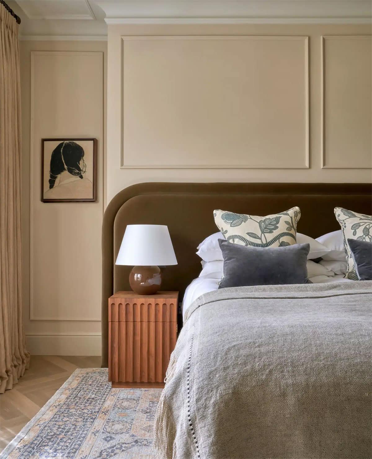 A cozy bedroom with a brown upholstered headboard, beige bedding, patterned pillows, a wooden nightstand with a white lamp, a framed abstract artwork on the wall, and a neutral rug on wooden floors.