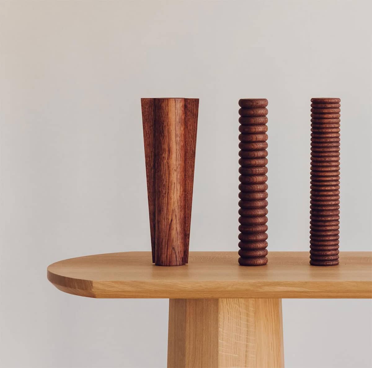 Three wooden sculptures sit on a light wood surface: one tall, smooth cone-shaped piece and two shorter, cylindrical pieces with ridged textures, all against a plain, light background.