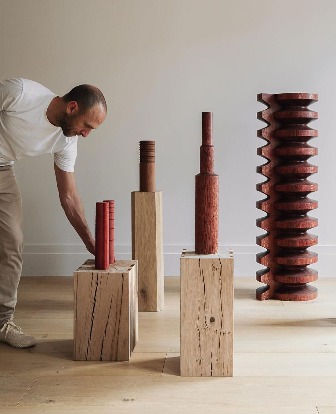 A person arranges modern wooden sculptures of varying heights and shapes on wooden plinths in a minimalist room with light-colored walls and flooring.
