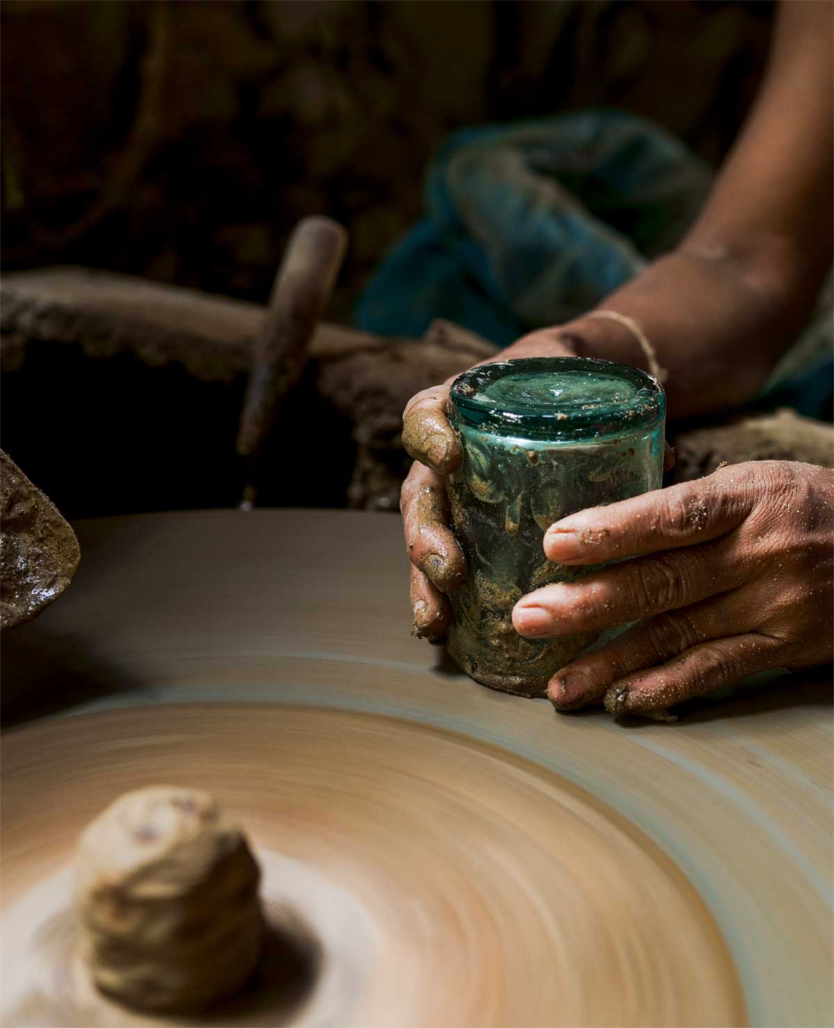 A person with clay-covered hands shapes a small green glass on a spinning pottery wheel, with blurred clay in the foreground and soft lighting highlighting the hands.