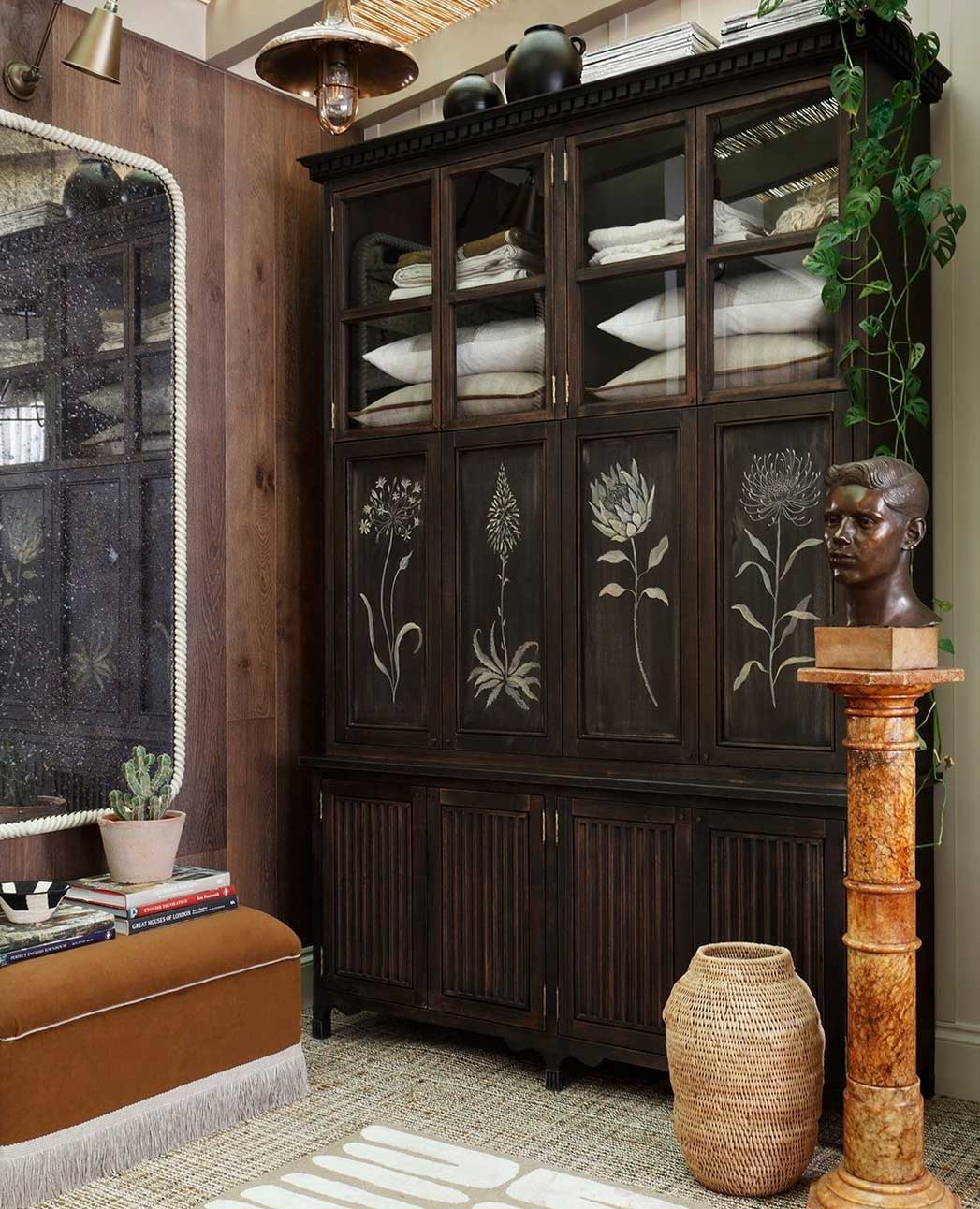 A dark wooden cabinet with etched floral designs holds towels. Next to it are a tall pedestal with a sculpted bust, a large vase, a brown bench, potted plants, and a mirror reflecting the room’s decor.