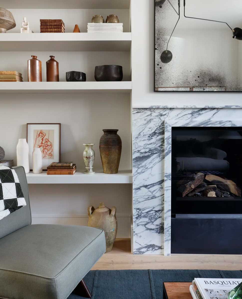 A modern living room with open shelves holding vases, books, and decor, a marble fireplace, abstract artwork above, and part of a gray armchair and coffee table in the foreground.