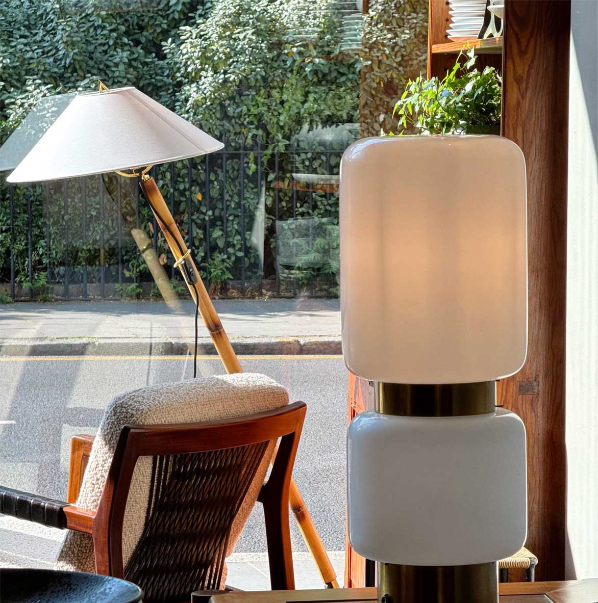 A modern table lamp with two white, rounded rectangular shades sits indoors near a window. Outside, a wicker chair and a standing lamp with a white shade are visible by the street and greenery.