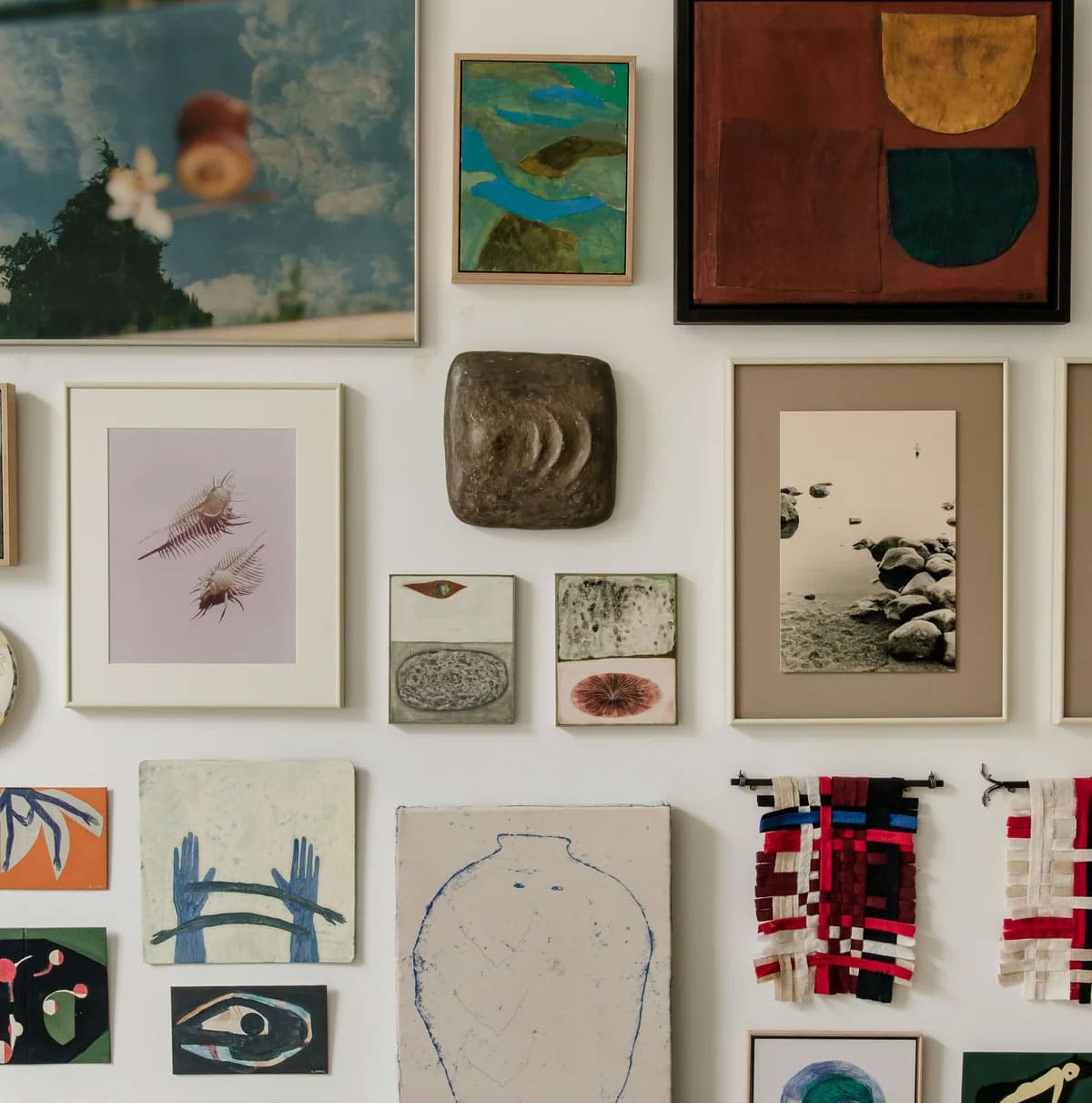 A gallery wall with various artworks including abstract paintings, photographs, textured pieces, and woven fabric art, all framed in different styles and sizes, displayed on a white wall.