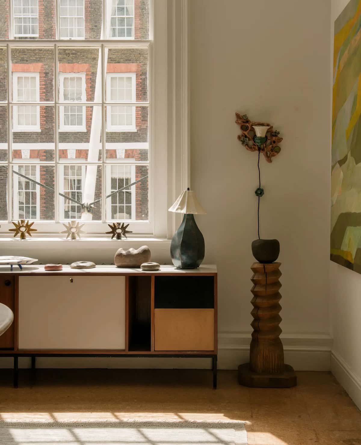 A sunlit room with a large window, a sideboard displaying decor items, a unique wooden floor lamp, and part of a colorful abstract painting on the wall. Shadows from the window fall across the wooden floor.