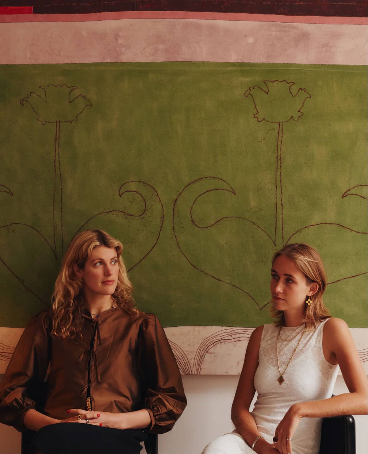 Two women sit side by side in front of a green wall tapestry with abstract floral designs. One wears a brown blouse with puffy sleeves, and the other wears a sleeveless white top. They both have long, wavy hair.