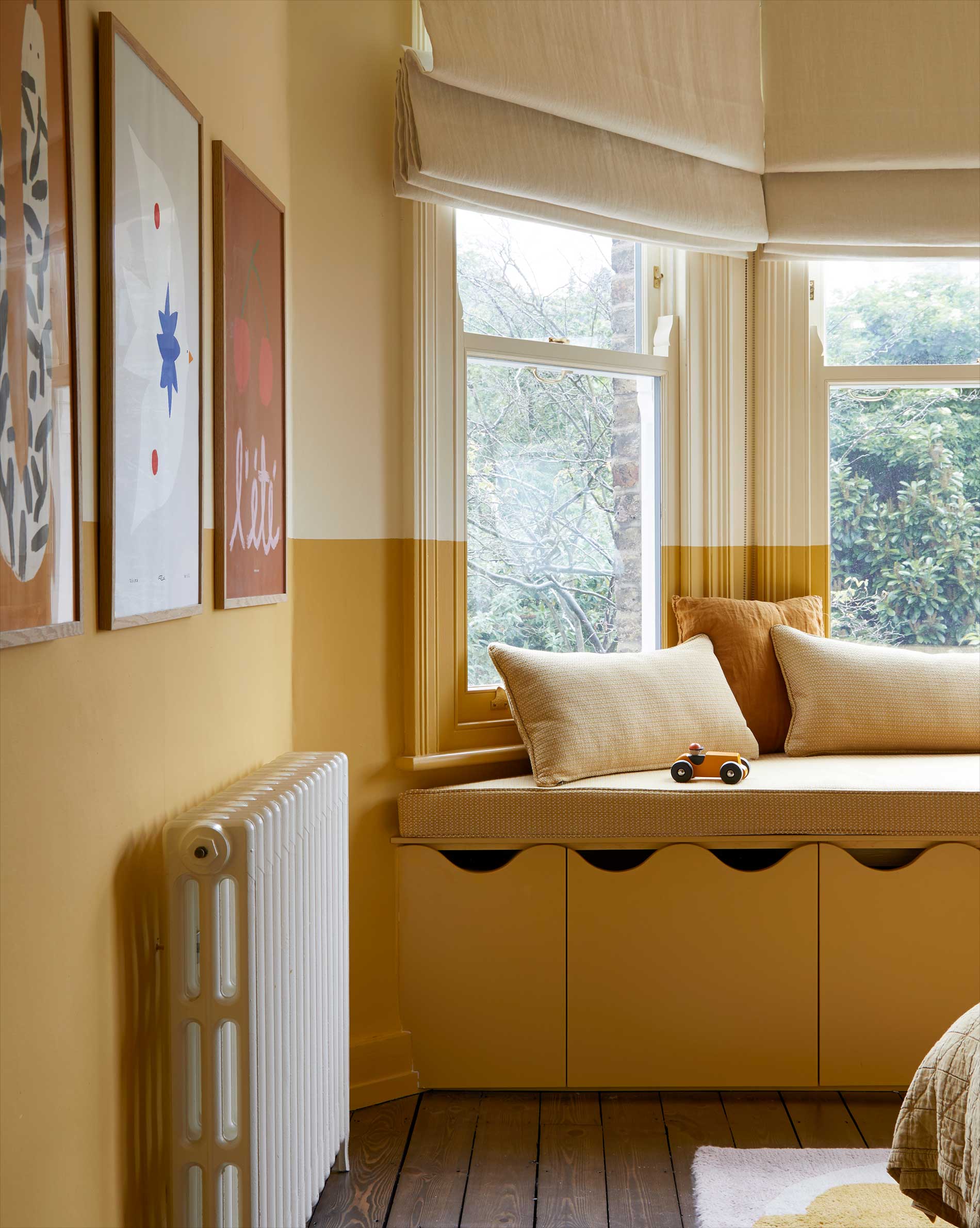 A cozy window nook with yellow walls, beige cushions, and a small toy car on the seat. Large windows bring in natural light, and framed artwork decorates the wall above a white radiator.