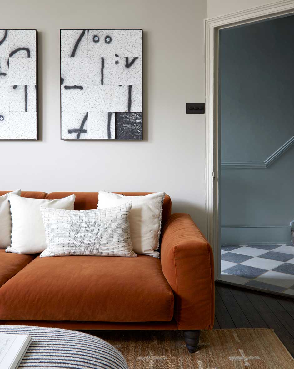 A rust-colored sofa with white and patterned cushions sits against a light gray wall, beneath two abstract black-and-white artworks. An open doorway reveals a tiled floor and a staircase beyond.