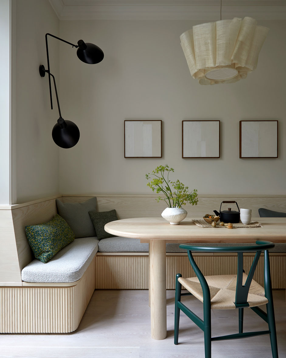 A modern dining nook features a light wood table, a built-in bench with cushions, a woven chair, minimal wall art, a black wall lamp, and a neutral pendant light, with a plant and tea set on the table.