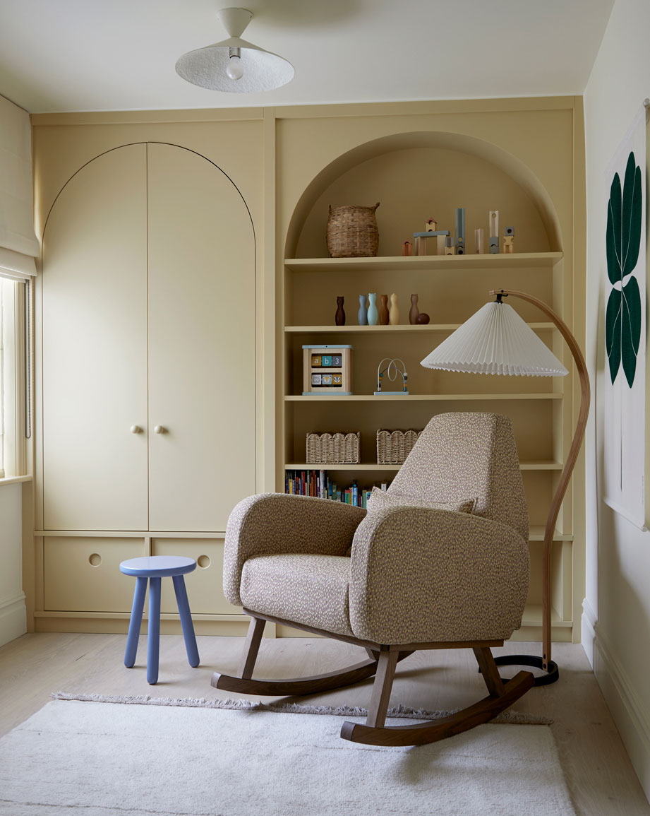 A cozy nursery features a beige rocking chair with wooden legs, a blue stool, a white rug, and a large arched built-in shelf filled with books, baskets, and decor. Soft natural light enters through a window on the left.