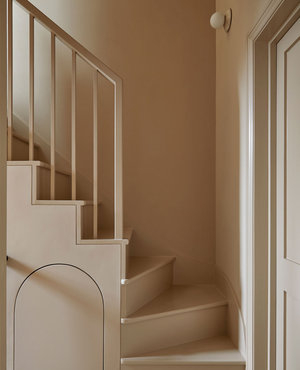 A beige-painted staircase with simple vertical railings, clean lines, and a curved doorway detail beneath the steps. The walls, stairs, and door are all a matching warm neutral color, creating a minimalist, modern look.