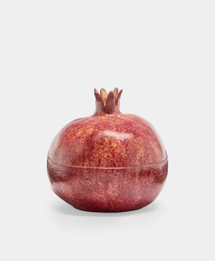 A ceramic container shaped and colored like a pomegranate, featuring a round body with a textured red surface and a crown-like top on a plain white background.