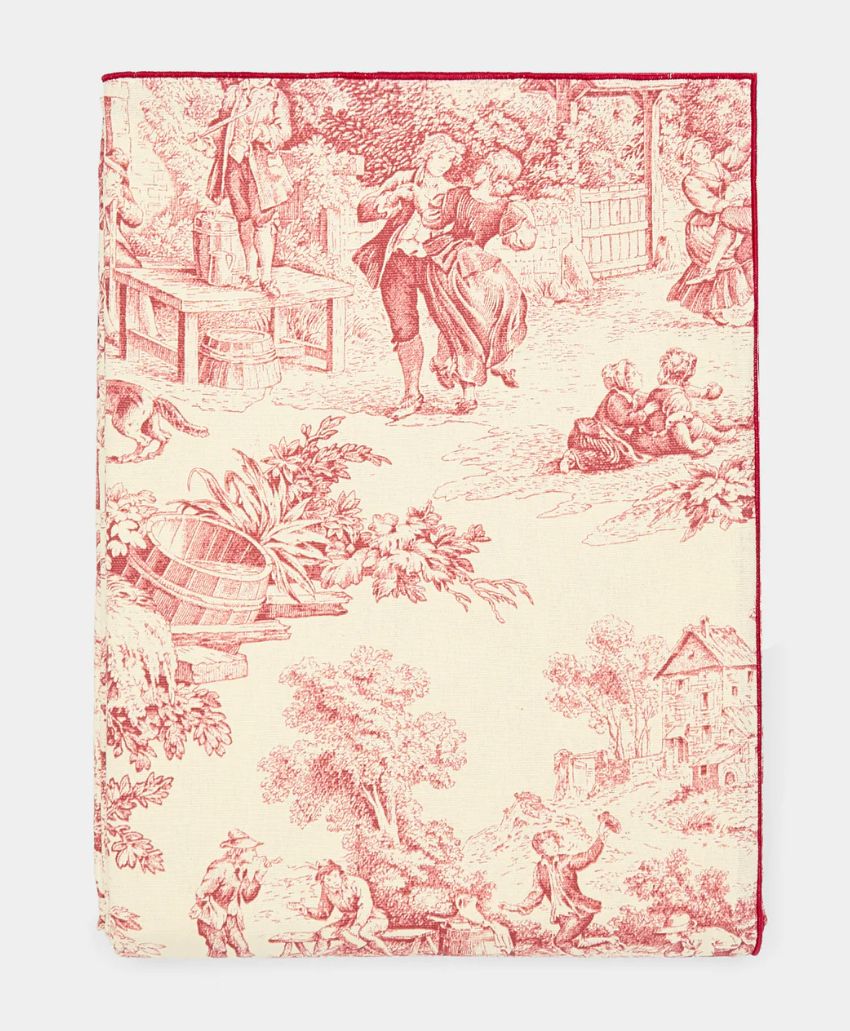 A fabric with red pastoral toile print showing people dancing, children playing, and rural scenery with trees, a house, and baskets, all on a cream background.