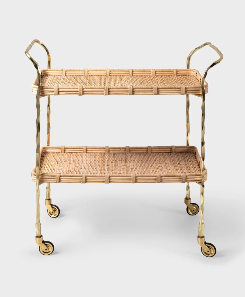 A two-tiered wicker and brass bar cart with basket-woven shelves, curved handles, and four swivel caster wheels, set against a plain light background.