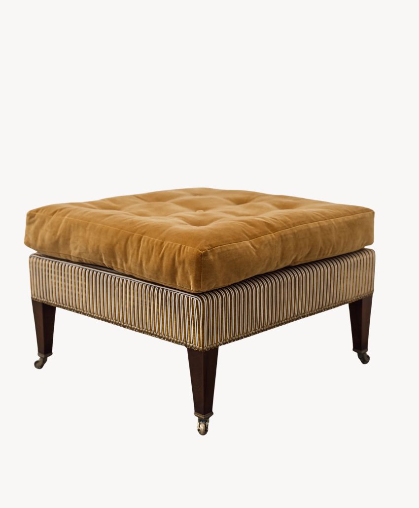 A square ottoman with a tufted golden-brown velvet cushion on top and a striped fabric base, supported by four dark wooden legs, two of which have small wheels.