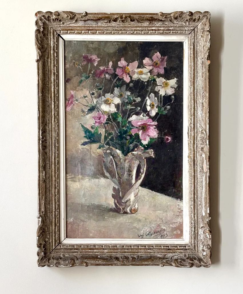 A framed painting of a white vase holding pink and white flowers sits on a light-colored surface against a dark background. The ornate frame has carved detailing.