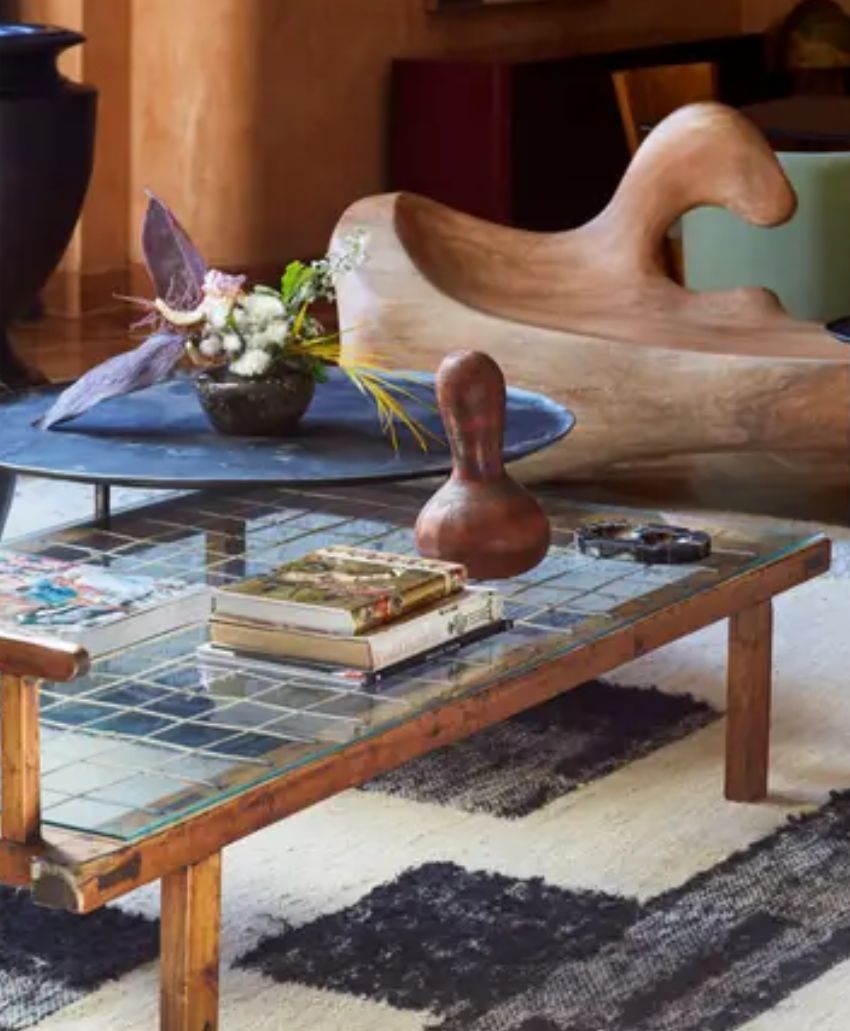 A coffee table with a tiled top holds books, a sculptural object, and small items. Behind it, a low round table displays a flower arrangement, and a wooden abstract chair is visible in the background.