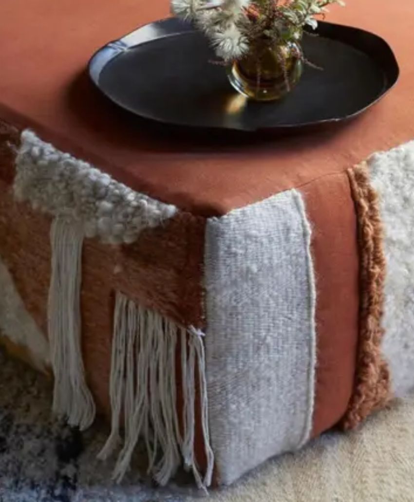A close-up of a textured ottoman with brown and cream woven fabric. A black tray holding a small vase with white flowers sits on top. Long cream yarn fringe hangs from the side of the ottoman.