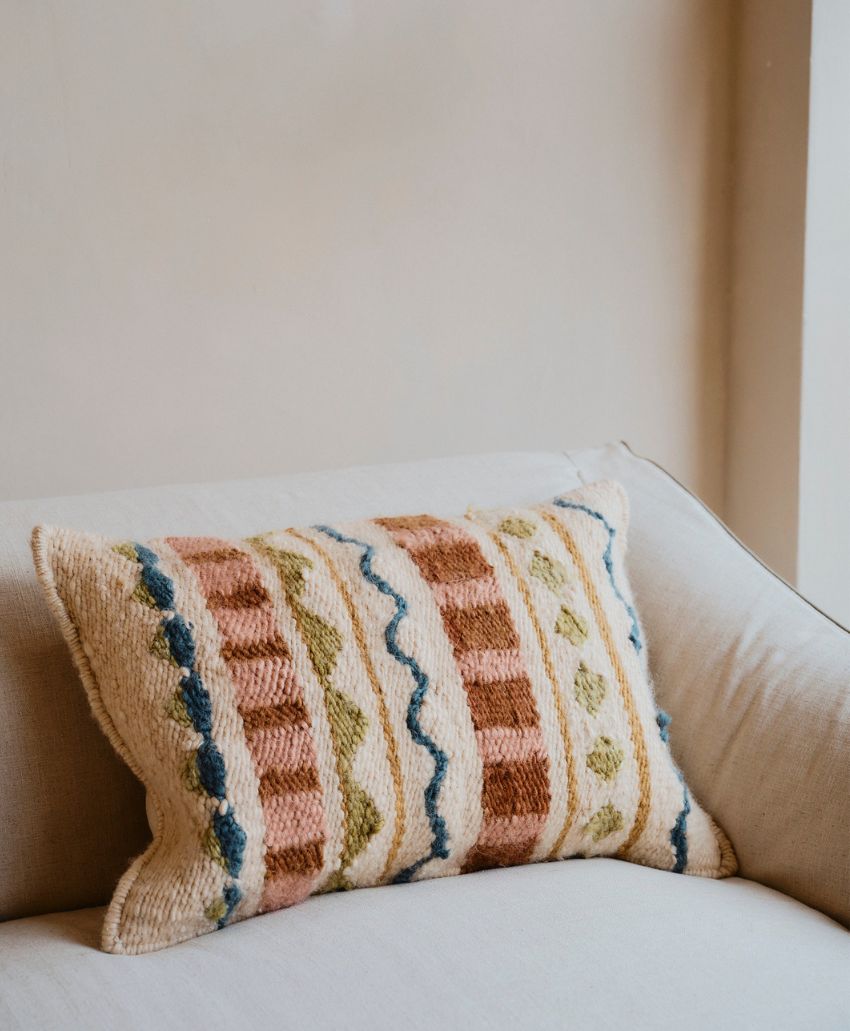 A decorative rectangular pillow with textured patterns in blue, brown, and green lies on a light beige sofa against a plain, light-colored wall.