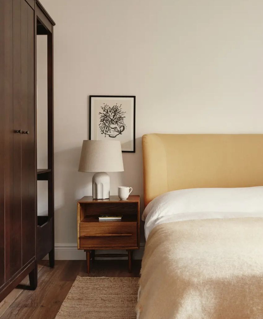 A cozy bedroom with a yellow upholstered bed, a wooden nightstand holding a lamp and a cup, a framed botanical print on the wall, and a dark wooden wardrobe beside a textured rug on the floor.