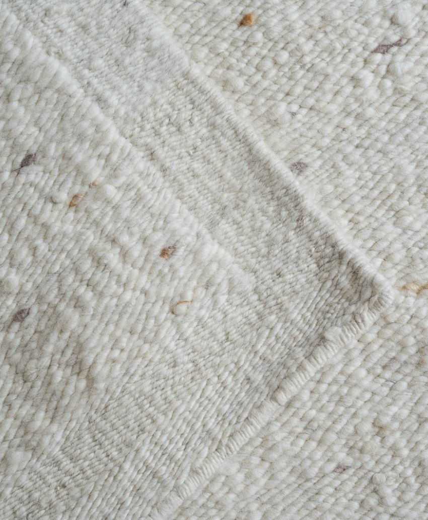 A close-up view of a textured, off-white woven rug with subtle beige and brown specks and visible overlap of its corners. The fabric appears soft and thick, highlighting the weave pattern and natural fibers.