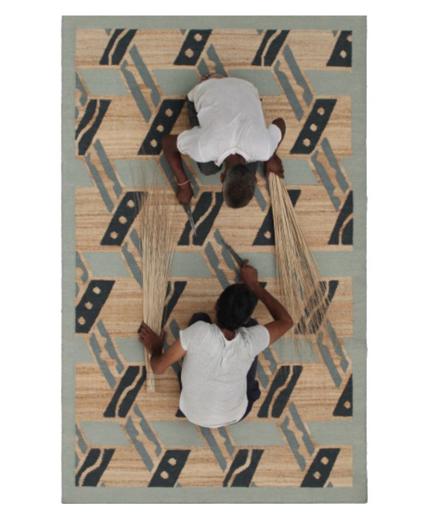 Close-up of a woven tabletop with a wooden tray holding two small ceramic bowls, next to a textured, cream-colored woven rug with tassels. Only the corner of the table and part of the rug are visible.