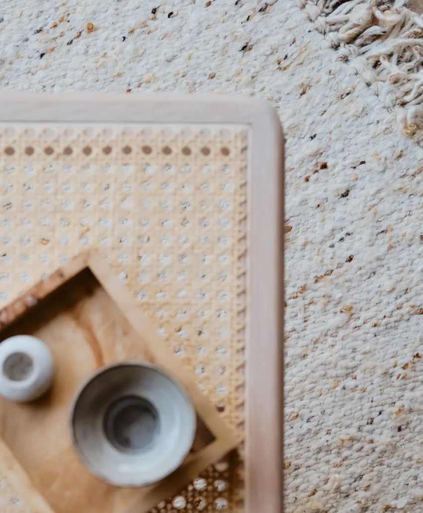 Close-up of a woven tabletop with a wooden tray holding two small ceramic bowls, next to a textured, cream-colored woven rug with tassels. Only the corner of the table and part of the rug are visible.