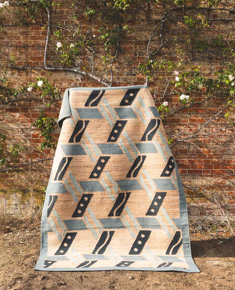 A patterned rug with geometric shapes and earthy tones is draped over a stand in front of a brick wall covered with leafy vines and white flowers.