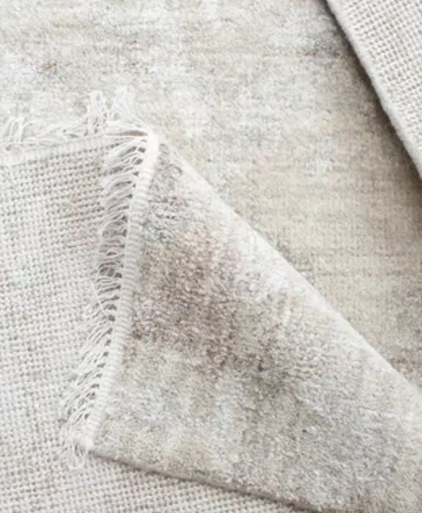 A close-up of a light beige woven rug, showing its textured surface and fringed edge with one corner folded over to reveal the underside.