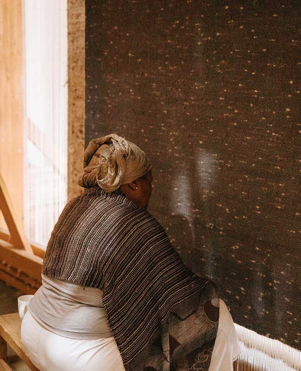 A woman wearing a headscarf and a shawl sits on a wooden bench, facing a large, dark, textured textile in a warm, softly lit room.