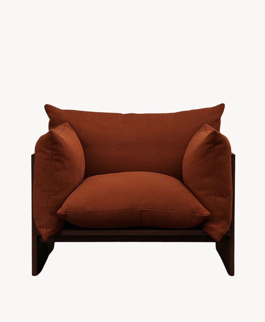 A modern armchair with a dark wooden frame, featuring thick, rust-colored cushions on the seat, back, and sides, set against a plain white background.