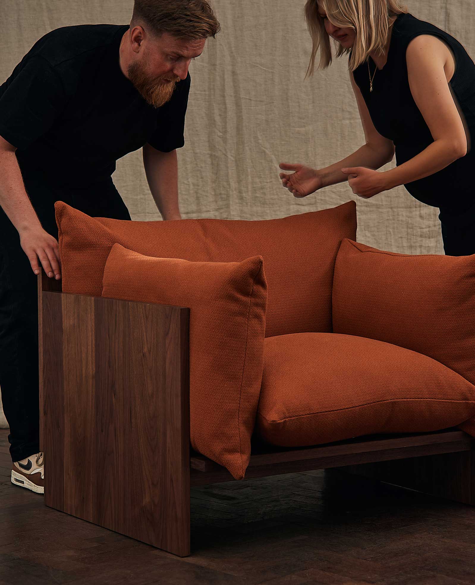 Two people position a large, modern armchair with wooden sides and plush, rust-colored cushions on a dark floor, against a beige fabric backdrop.