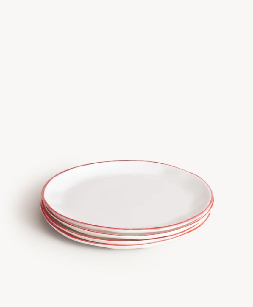 A stack of four white ceramic plates with thin red rims, placed on a white surface against a plain white background.