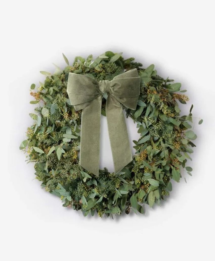 A green wreath made of assorted foliage, decorated with a large, soft green velvet bow at the top, displayed against a plain white background.