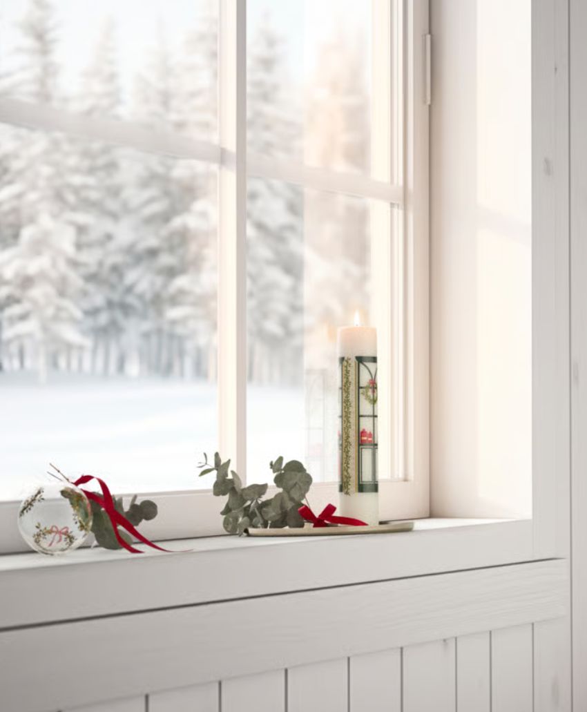 A white candle, greenery, and a Christmas ornament with a red ribbon decorate a windowsill. Outside the window, snow-covered trees and a wintry landscape are visible.