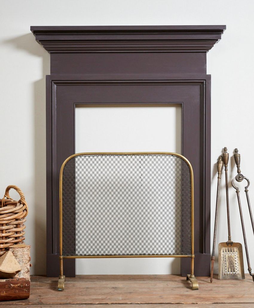 A dark wood fireplace surround with a brass mesh firescreen in front, a basket of firewood on the left, and three brass fireplace tools on the right, all set against a light-colored wall and wooden floor.