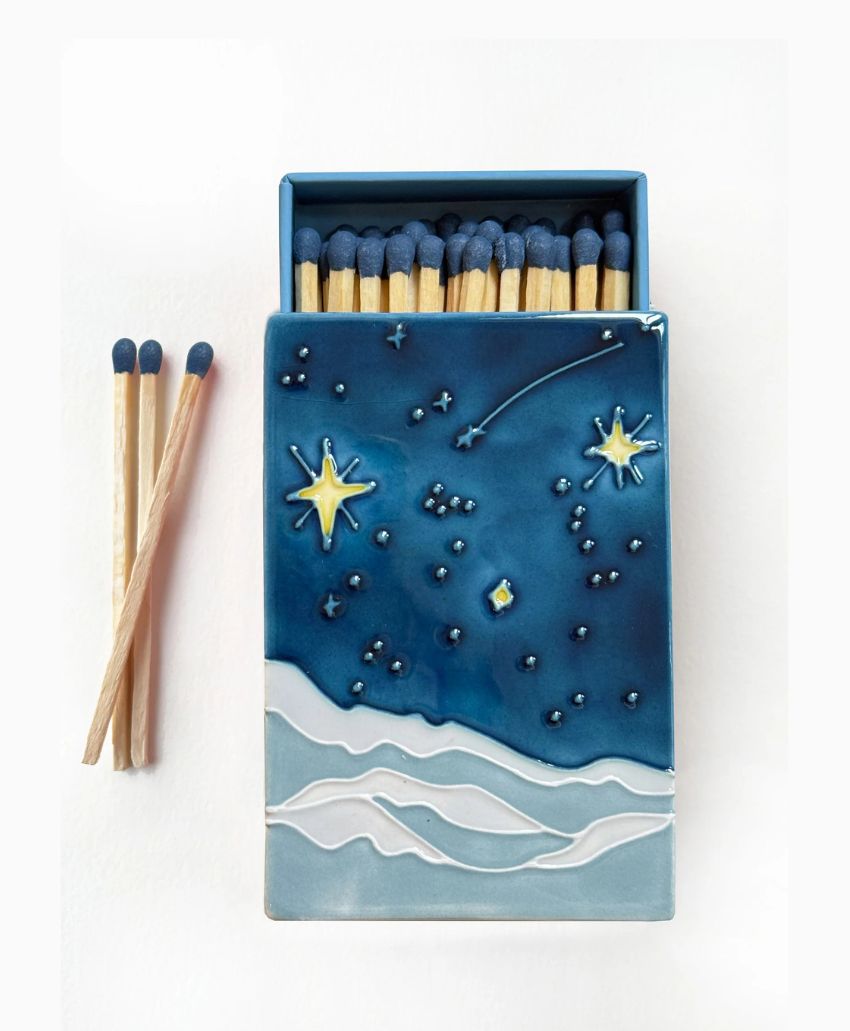 A box of matches with an illustrated cover showing a starry night sky, constellations, and shooting star above white, wavy hills. Three matches are placed next to the box on a white background.