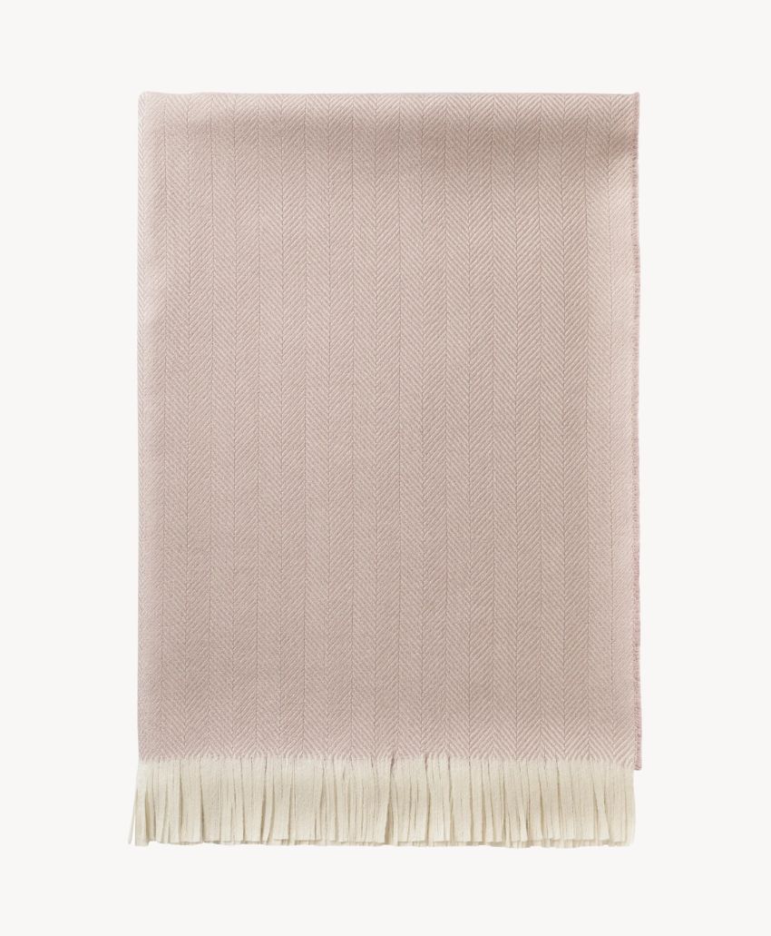 A folded, light pink herringbone-patterned throw blanket with cream-colored fringe along the bottom edge, displayed on a plain, light background.