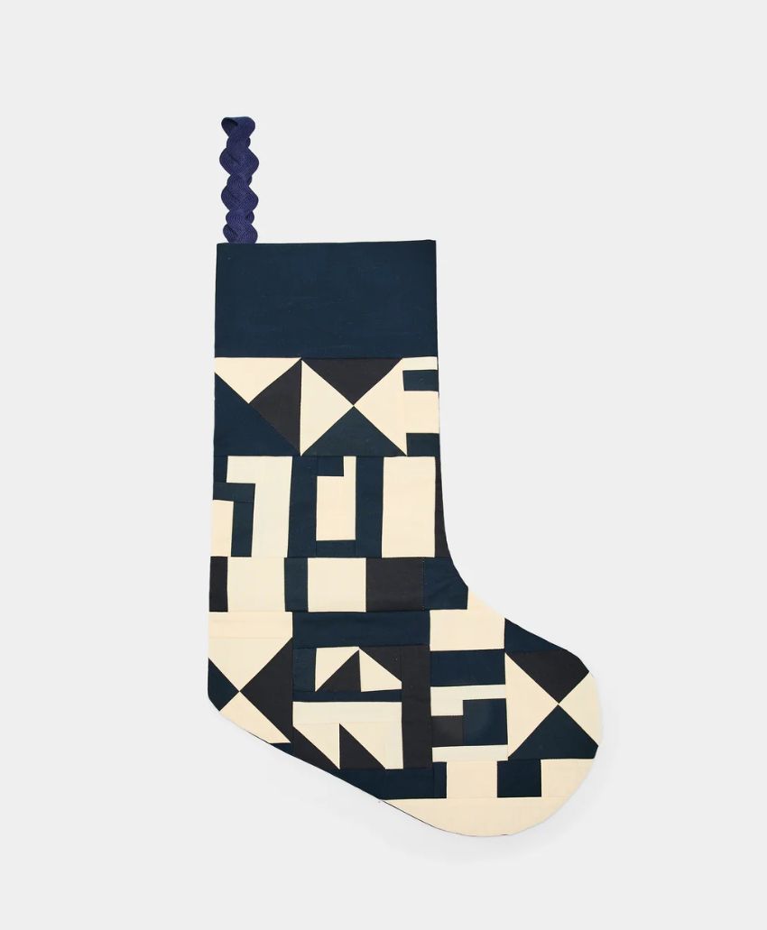 A Christmas stocking with a dark blue and cream geometric patchwork pattern and a braided blue loop for hanging, displayed on a plain light background.