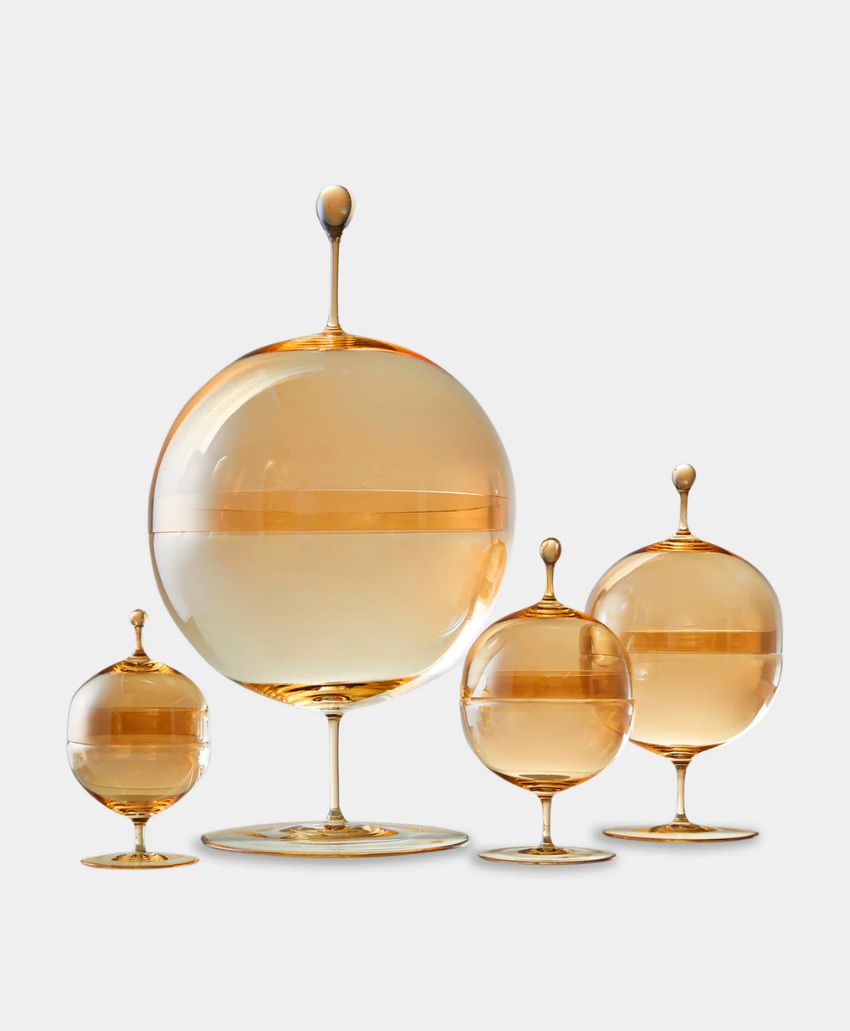 Four amber-colored glass globes of varying sizes, each on a thin stem and flat base, are arranged together against a plain white background. The globes have round tops and reflective surfaces.