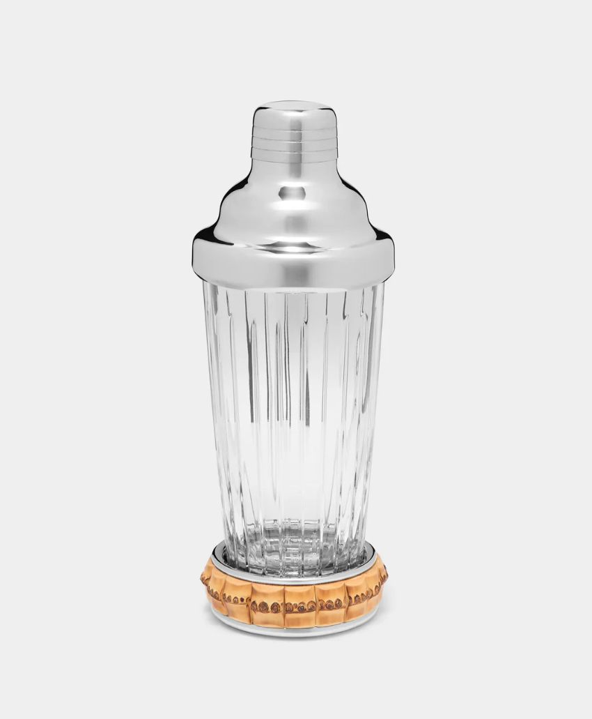 A clear glass cocktail shaker with vertical ridges, a shiny silver metal lid, and a decorative gold band around the base, displayed against a plain light gray background.