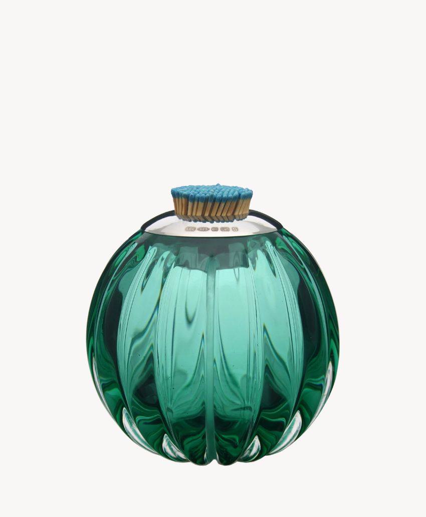 A round, green glass match striker with vertical ridges, filled with blue-tipped matches standing upright on top of a white striker pad, set against a plain white background.