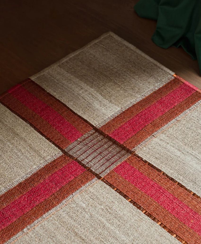 A beige woven rug with intersecting red and brown stripes forming a cross pattern, placed on a brown wooden floor with a green fabric partially visible in the corner.